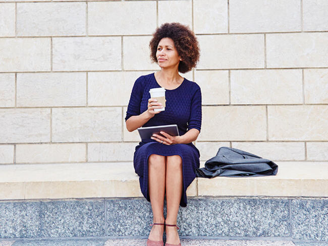 A woman sits on a step outdoors drinking coffee, working on tablet device